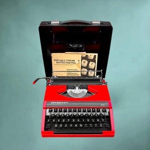 Könnte beinhalten: Eine leuchtend rote tragbare Schreibmaschine Sperry Remington 1010 mit schwarzem Kofferdeckel. Das Gerät verfügt über eine schwarze Tastatur mit AZERTY-Layout und einen eingesteckten Papier-Anleitungsordner mit der Aufschrift Portable Typewriter Instruction Folder.