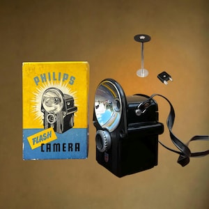 Peut inclure: Un appareil photo flash Philips noir avec un réflecteur argenté, une sangle noire, un petit support en métal et une prise à deux broches. À côté se trouve une boîte en carton jaune et bleue avec le texte Philips Flash Camera et une illustration.
