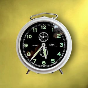 Könnte beinhalten: Ein weißer Vintage-Wecker mit schwarzem Zifferblatt und leuchtend grünen Ziffern. Die Uhr verfügt über eine Wiederholungsfunktion und eine Alarmeinstellung, mit einem Metallgriff und Beinen. Das Zifferblatt zeigt die Wörter "REPEAT" und "ALARM".