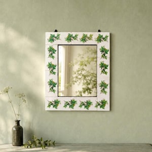 May include: A rectangular wall mirror featuring a frame constructed from white ceramic tiles. Each tile is decorated with a green ivy leaf pattern. The mirror has a central reflective glass panel and two black metal mounting brackets at the top.