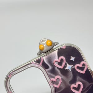 May include: A clear phone case featuring a dark, wavy background pattern decorated with light pink hand-drawn hearts and small white star icons. A small, three-dimensional gray figure with two bright orange circular eyes sits on the top edge.