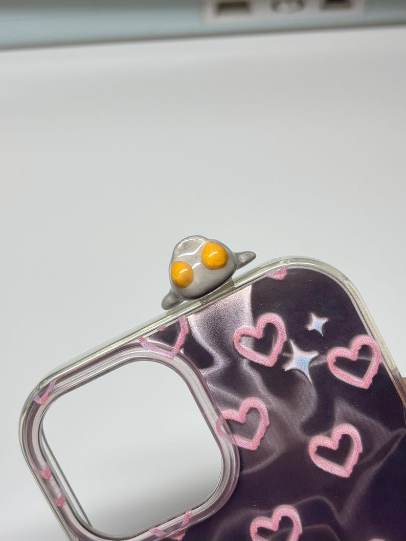 May include: A clear phone case featuring a dark, wavy background pattern decorated with light pink hand-drawn hearts and small white star icons. A small, three-dimensional gray figure with two bright orange circular eyes sits on the top edge.