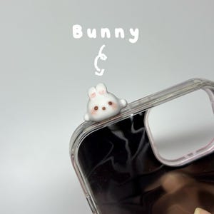 May include: A clear phone case featuring a small, white, three-dimensional bunny figurine attached to the top corner. The bunny has pink-tinted ears, dark eyes, and a brown nose. The word Bunny is written in white text above the figurine.