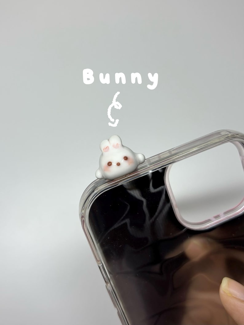 May include: A clear phone case featuring a small, white, three-dimensional bunny figurine attached to the top corner. The bunny has pink-tinted ears, dark eyes, and a brown nose. The word Bunny is written in white text above the figurine.