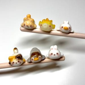May include: Six small, glossy, animal-shaped ceramic figures are perched on two light-colored wooden chopsticks. The figures include a calico cat, a yellow pufferfish, a white duck, a brown and white cat, a grey penguin, and a white rabbit.