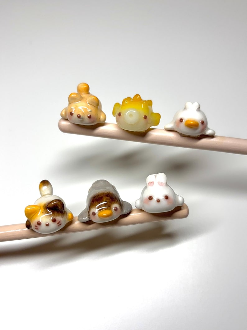 May include: Six small, glossy, animal-shaped ceramic figures are perched on two light-colored wooden chopsticks. The figures include a calico cat, a yellow pufferfish, a white duck, a brown and white cat, a grey penguin, and a white rabbit.
