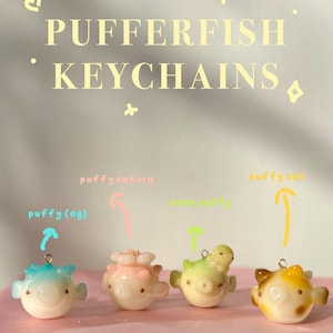 May include: Four small pufferfish keychains with metal loops. The first is blue and white, the second is pink with a flower, the third is green with a small octopus, and the fourth is yellow and brown. Text reads PUFFERFISH KEYCHAINS, puffy(og), puffy sakura, octopuffy, and puffy cat.