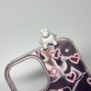 May include: A clear phone case featuring a dark, wavy, metallic-looking background pattern decorated with small, hand-drawn style pink hearts and tiny white stars. A small, white, glossy, three-dimensional figure of a creature is attached to the top.