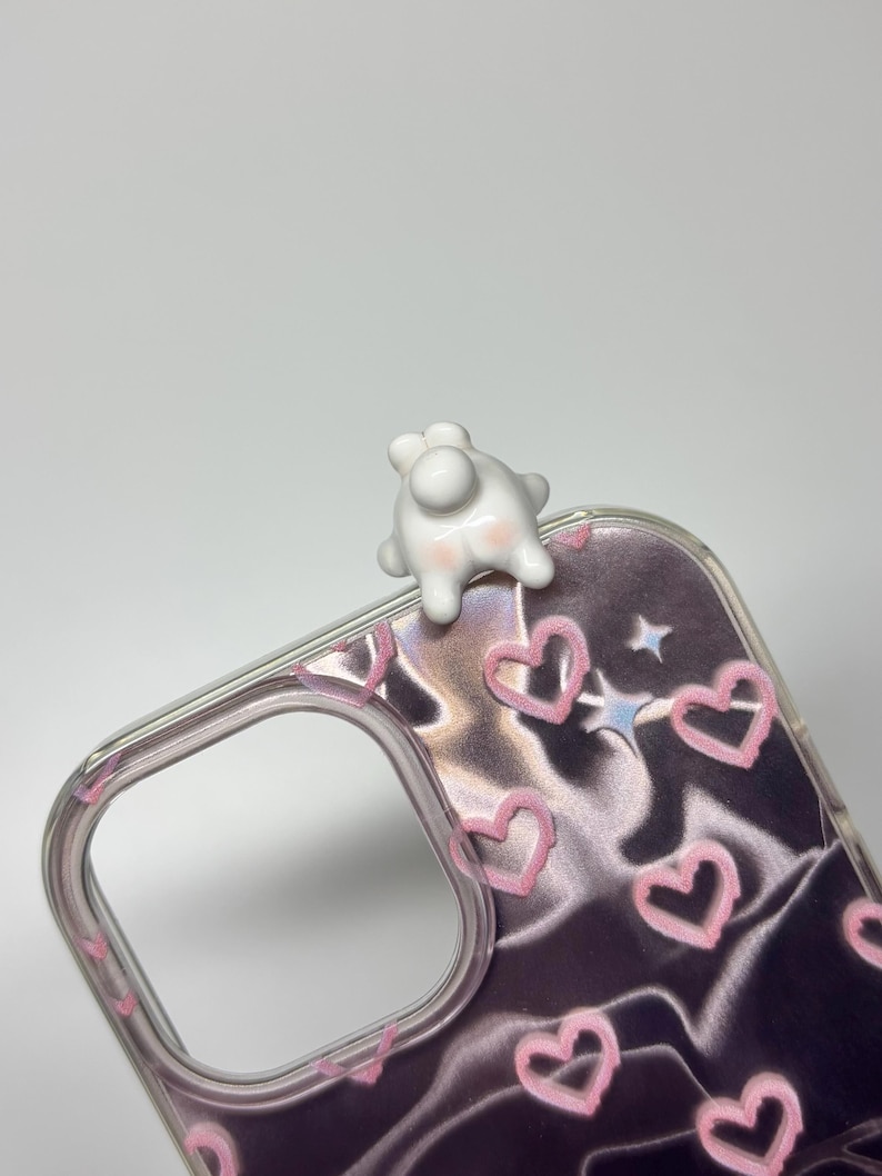 May include: A clear phone case featuring a dark, wavy, metallic-looking background pattern decorated with small, hand-drawn style pink hearts and tiny white stars. A small, white, glossy, three-dimensional figure of a creature is attached to the top.