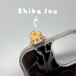 May include: A clear phone case featuring a small, three-dimensional ceramic Shiba Inu dog figurine attached to the top corner. The text "Shiba Inu" is printed in white above the figurine, accompanied by a small, hand-drawn white swirl graphic.