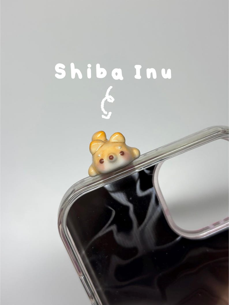 May include: A clear phone case featuring a small, three-dimensional ceramic Shiba Inu dog figurine attached to the top corner. The text "Shiba Inu" is printed in white above the figurine, accompanied by a small, hand-drawn white swirl graphic.