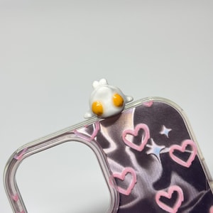 May include: A clear phone case featuring a dark, satin-like background with a pattern of light pink hand-drawn hearts and small blue stars. A small, white, three-dimensional character with two bright orange circular eyes sits on the top edge.