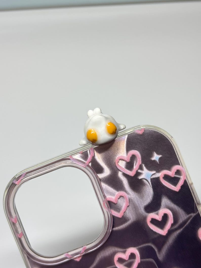 May include: A clear phone case featuring a dark, satin-like background with a pattern of light pink hand-drawn hearts and small blue stars. A small, white, three-dimensional character with two bright orange circular eyes sits on the top edge.