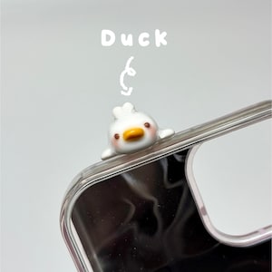 May include: A clear phone case featuring a small, white, three-dimensional duck figurine attached to the top edge. The duck has a yellow beak, brown eyes, and pink cheeks. The word Duck is printed in white above the duck with a hand-drawn arrow.
