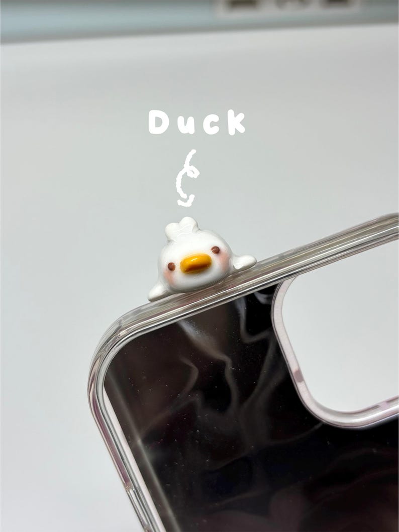 May include: A clear phone case featuring a small, white, three-dimensional duck figurine attached to the top edge. The duck has a yellow beak, brown eyes, and pink cheeks. The word Duck is printed in white above the duck with a hand-drawn arrow.