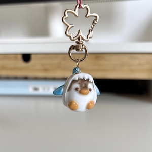 Puede incluir: Un pequeño llavero con un charm de pingüino blanco, marrón y azul. El pingüino tiene patas y pico naranjas. El llavero presenta un cierre en forma de flor dorada.