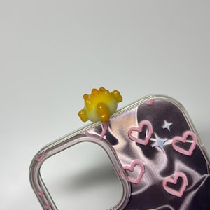 May include: A clear phone case featuring a dark, metallic, wavy background pattern with pink hand-drawn heart outlines and small star shapes. A small, three-dimensional yellow and orange glass dinosaur figure is attached to the top edge.