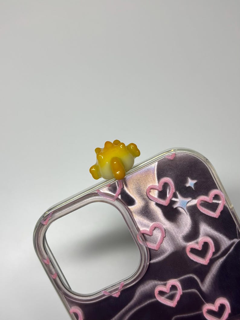 May include: A clear phone case featuring a dark, metallic, wavy background pattern with pink hand-drawn heart outlines and small star shapes. A small, three-dimensional yellow and orange glass dinosaur figure is attached to the top edge.