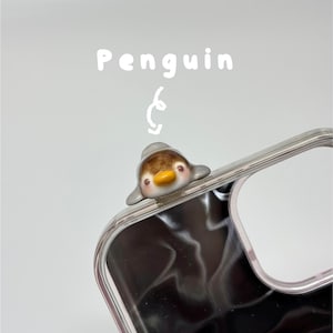 May include: A clear phone case featuring a small, three-dimensional ceramic penguin figurine attached to the top corner. The penguin has a brown head, grey wings, and a yellow beak. Above the penguin, the word Penguin is written in white text.