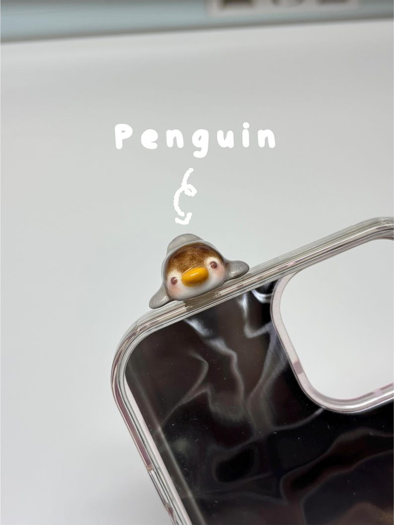 May include: A clear phone case featuring a small, three-dimensional ceramic penguin figurine attached to the top corner. The penguin has a brown head, grey wings, and a yellow beak. Above the penguin, the word Penguin is written in white text.