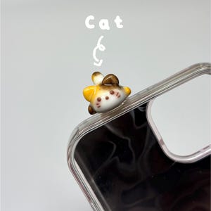 May include: A clear phone case featuring a small, three-dimensional ceramic cat figurine attached to the top corner. The cat is white, brown, and yellow with painted facial features. The word Cat is printed in white above the figurine.