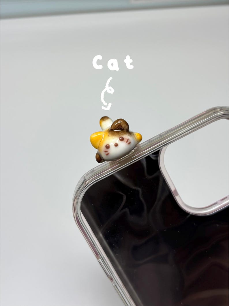 May include: A clear phone case featuring a small, three-dimensional ceramic cat figurine attached to the top corner. The cat is white, brown, and yellow with painted facial features. The word Cat is printed in white above the figurine.
