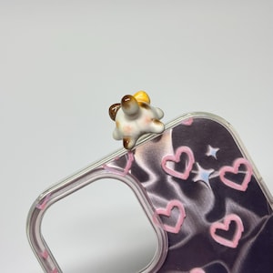 May include: A clear phone case featuring a dark purple background with a pattern of pink hand-drawn hearts and small white stars. Attached to the top edge of the case is a small, three-dimensional ceramic figurine of a calico cat's rear end.