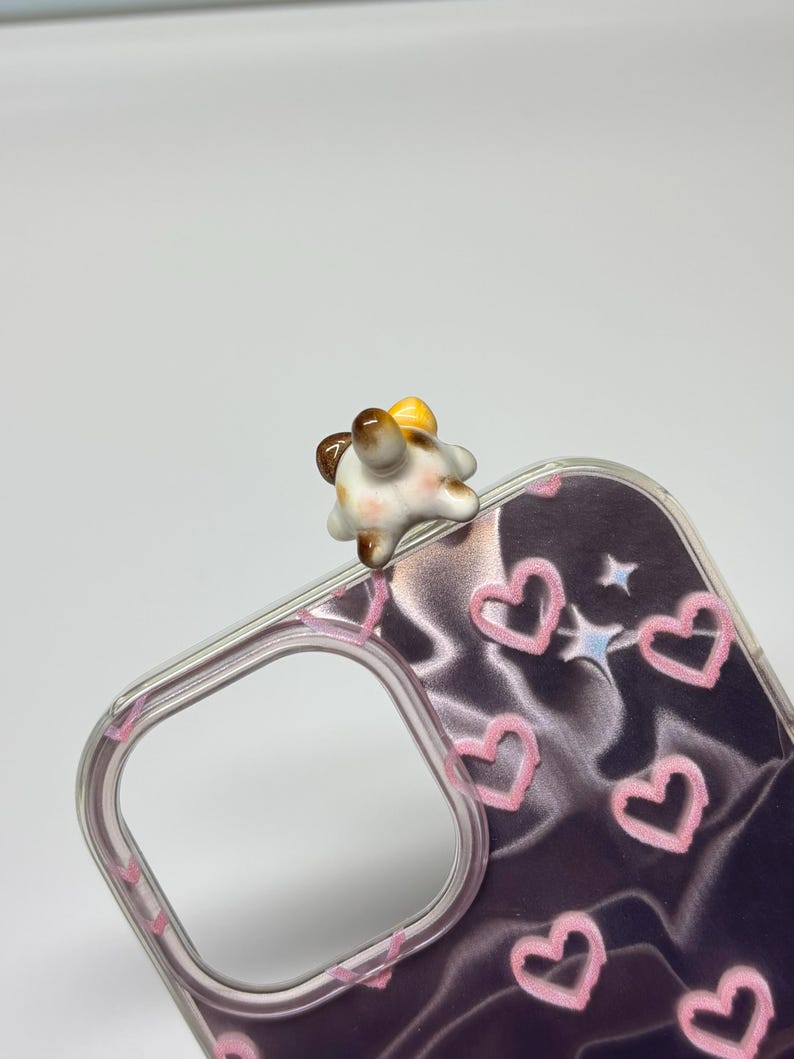 May include: A clear phone case featuring a dark purple background with a pattern of pink hand-drawn hearts and small white stars. Attached to the top edge of the case is a small, three-dimensional ceramic figurine of a calico cat's rear end.