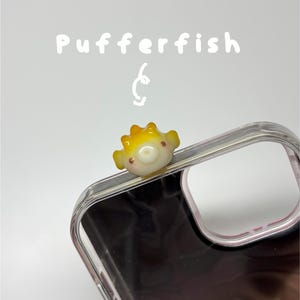 May include: A clear phone case featuring a small, three-dimensional yellow and white pufferfish charm attached to the top edge. The text Pufferfish is printed in white above the charm, accompanied by a small white hand-drawn swirl graphic.