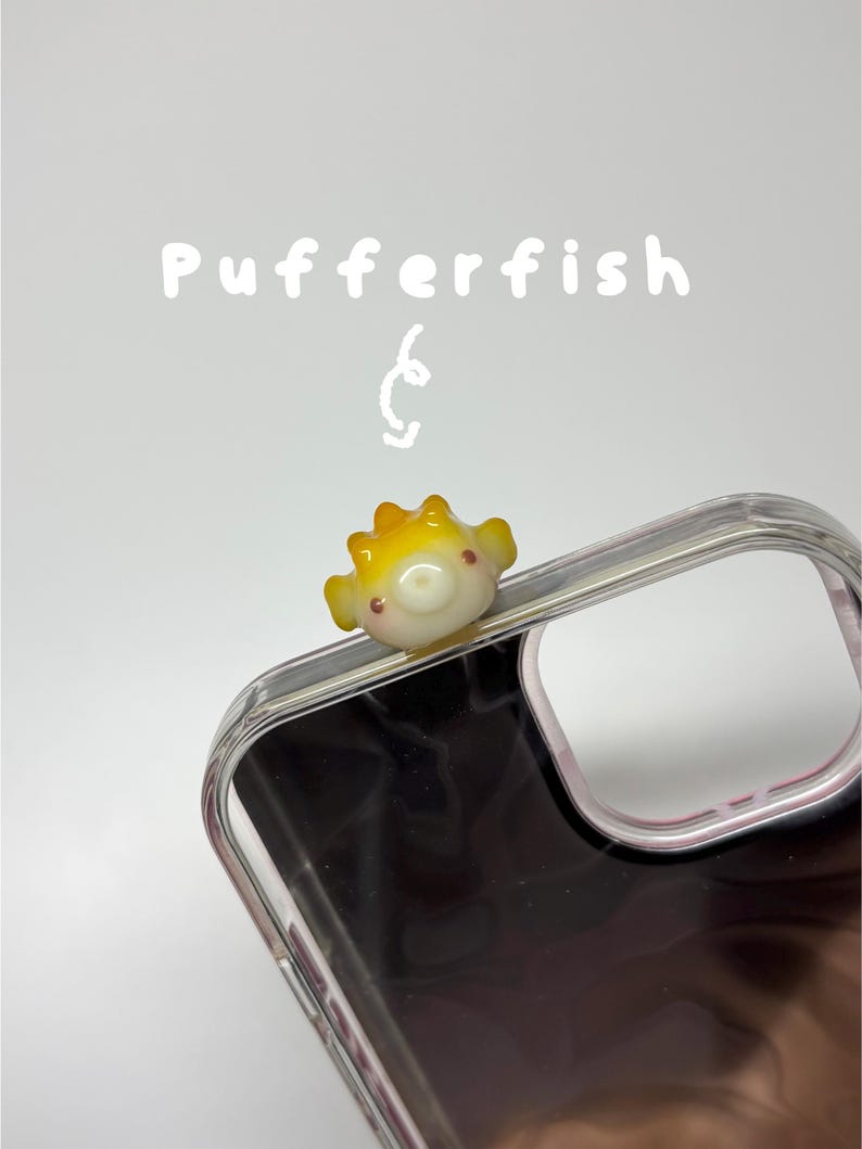 May include: A clear phone case featuring a small, three-dimensional yellow and white pufferfish charm attached to the top edge. The text Pufferfish is printed in white above the charm, accompanied by a small white hand-drawn swirl graphic.