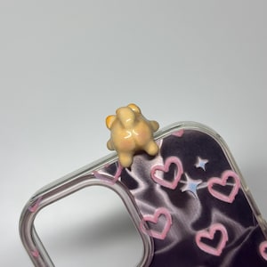 May include: A clear phone case featuring a dark purple background with a pattern of light pink hand-drawn hearts and small white stars. A small, three-dimensional, beige-colored animal figurine is attached to the top edge of the case.