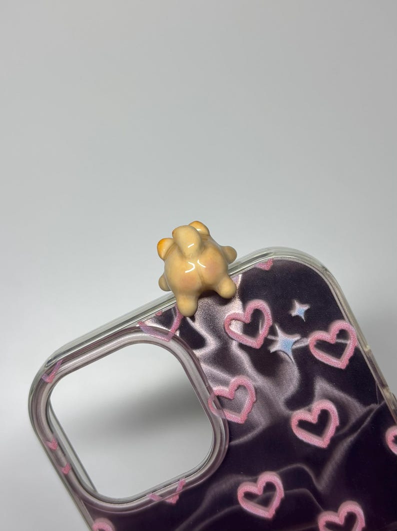 May include: A clear phone case featuring a dark purple background with a pattern of light pink hand-drawn hearts and small white stars. A small, three-dimensional, beige-colored animal figurine is attached to the top edge of the case.