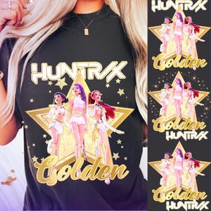 Puede incluir: Camiseta negra con un diseño de estrella dorada que presenta tres figuras animadas con trajes blancos. El texto "HUNTRAX" está encima de la estrella y "Golden" debajo. Pequeñas estrellas están dispersas alrededor del diseño.