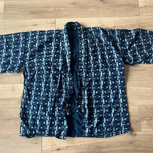Puede incluir: Una chaqueta haori con estampado azul oscuro y blanco que presenta un diseño geométrico repetitivo de triángulos. La prenda tiene mangas anchas de tres cuartos, frente abierto y un forro interior azul oscuro sólido visible en el cuello.
