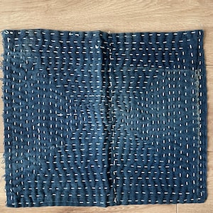 May include: A rectangular piece of indigo-dyed fabric featuring a dense pattern of white running stitches arranged in horizontal and vertical lines. The textile displays a weathered texture with frayed edges and a visible grid-like embroidery design.