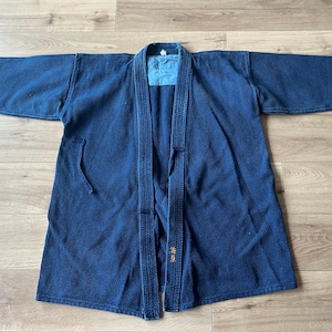 Op de afbeelding: Een donkerblauw indigo-geverfd kendo-jasje met een textuur van gewatteerde sashiko-stof, wijde mouwen en een verstevigde kraag. Het kledingstuk heeft een lichtblauw binnenvlak bij de nek en goudkleurig Japans kanji-borduurwerk.