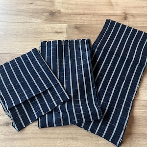 May include: Three rectangular pieces of dark navy blue fabric featuring a white pinstripe pattern with thin horizontal lines. The fabric pieces are arranged in varying sizes, showing raw, unfinished edges on all sides of the textile material.