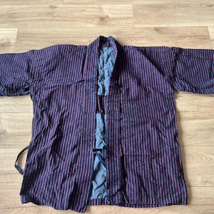 Peut inclure: Une veste haori à rayures bleu foncé et rouge dotée d'un large col, de manches trois-quarts et d'une fermeture à nouer sur le devant. Le vêtement présente une doublure bleu uni visible le long des bords intérieurs et une coupe ouverte.
