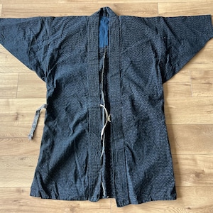 Könnte beinhalten: Eine dunkelblaue Jacke im japanischen Haori-Stil mit einem dezenten, fein linierten Gittermuster auf dem gesamten Stoff. Das Kleidungsstück hat weite, gerade Ärmel, einen strukturierten Kragen und ein einfarbig blaues Innenfutter mit dünnen Bindebändern.