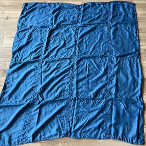 Puede incluir: Una manta rectangular de tela azul con un patrón de cuadrícula formado por costuras. El material tiene una textura ligeramente arrugada y un color azul profundo uniforme en toda la pieza, con líneas verticales y horizontales que crean secciones cuadradas.