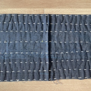 May include: A rectangular piece of dark charcoal grey fabric featuring horizontal rows of white running stitches. The textile has a visible quilted texture with a consistent pattern of dashed lines across the entire surface, creating a grid-like appearance.