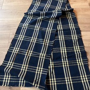 May include: A rectangular piece of fabric featuring a dark navy blue base colour with a repeating plaid pattern in light yellow and thin white lines. The textile has visible raw, unfinished edges along the bottom and sides of the material.
