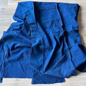 May include: A large piece of deep blue fabric with a visible woven texture. The material is draped in loose folds, showing raw, unfinished edges along the sides and bottom. The fabric has a matte finish and a consistent, solid dark blue colour.