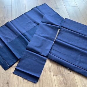 Puede incluir: Varias piezas de tela azul índigo se disponen sobre una superficie de madera. Las piezas de tela, de diversas formas y tamaños, parecen ser rectangulares o de forma irregular. La tela presenta un aspecto ligeramente texturizado.