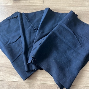 May include: A folded piece of dark navy blue fabric with a visible woven texture. The material is arranged in multiple layers, showing natural creases and folds across the surface of the textile, which appears to be a solid, opaque colour.