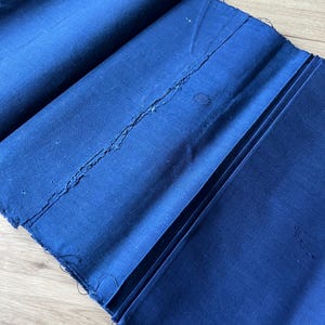Könnte beinhalten: Ein Stück tiefblauer Stoff, der in mehrere Abschnitte gefaltet ist. Das Material hat eine matte Textur mit sichtbaren, ausgefransten Kanten entlang der Nähte und einem kleinen kreisförmigen Fleck auf der Oberfläche. Der Stoff ist in Falten gelegt.