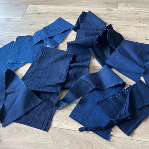 May include: A collection of multiple dark blue fabric strips with visible raw, frayed edges. The textile features a textured, woven appearance with varying shades of deep indigo and navy blue throughout the scattered pieces of material.
