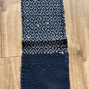Könnte beinhalten: Ein rechteckiges indigofarbenes Textil mit weißen Kreuzstichmustern im oberen Bereich und einfarbigem indigofarbenem Stoff im unteren Bereich. Die Kanten sind mit weißem Faden genäht. Das Textil liegt auf einer Holzoberfläche.