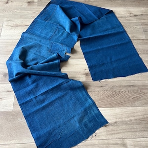 May include: A long, rectangular piece of fabric dyed in a deep indigo blue colour. The textile features a visible woven texture with raw, unfinished edges at both ends. The material is draped in a loose, flowing manner, showcasing variations in the blue hue.
