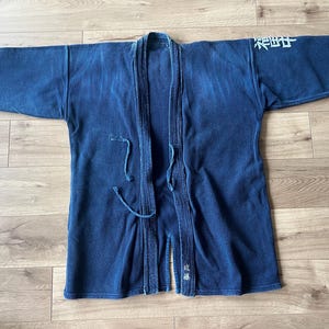 Könnte beinhalten: Eine dunkelblaue, indigogefärbte Kendo-Jacke mit texturiertem Sashiko-Gewebe, langen Ärmeln und einer Bindung auf der Vorderseite. Das Kleidungsstück zeigt weiße japanische Kanji-Schriftzeichen, die auf der linken Schulter und am Saum gestickt sind.
